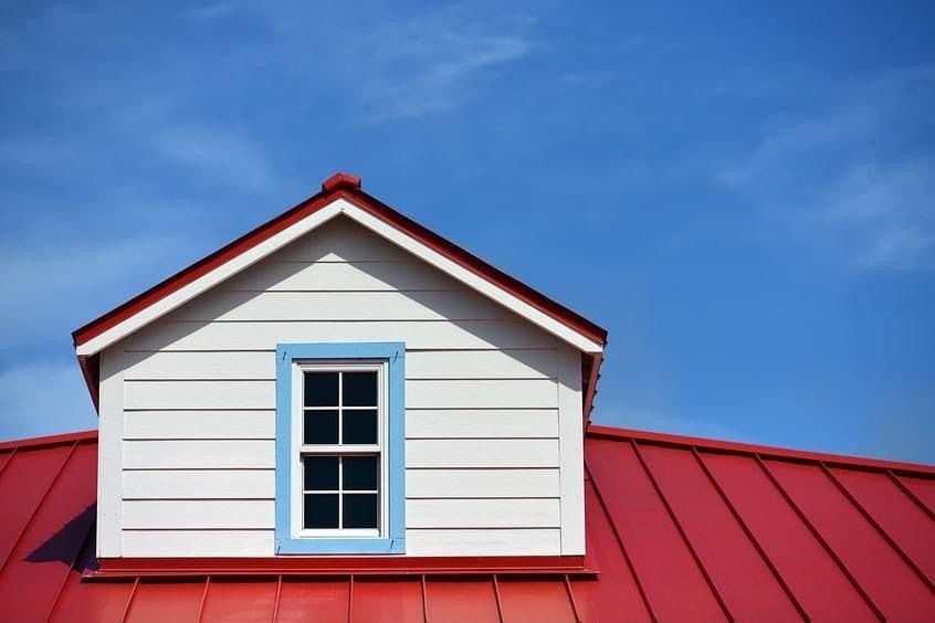 House with white siding, blue window trim, and red metal roof against a clear blue sky.